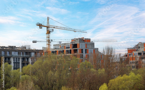 Obraz na płótnie A modern residential complex under construction, featuring concrete and brick structures with large glass windows, surrounded by trees, with cranes towering above against a clear blue sky. w salonie
