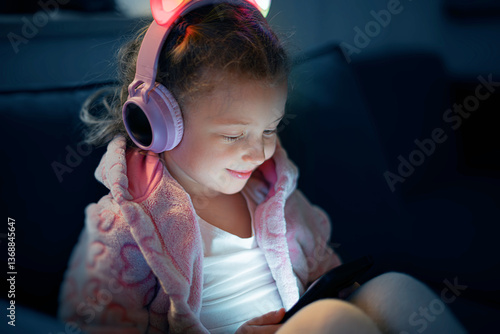 Obraz na płótnie A young girl wearing pink glowing cat-ear headphones and a cozy fleece jacket is happily engaged with a mobile device, her face illuminated by the screens soft light in a dimly lit room. w salonie