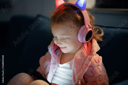 A young girl wearing pink glowing cat-ear headphones and a cozy fleece jacket is happily engaged with a mobile device, her face illuminated by the screens soft light in a dimly lit room.