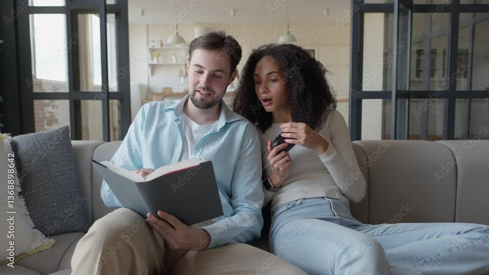 Couple Relaxing On Sofa, Reading Book Together, Reacting With Surprise Interest Couple Relaxing On Sofa, Reading Book Together, Reacting With Surprise Interest