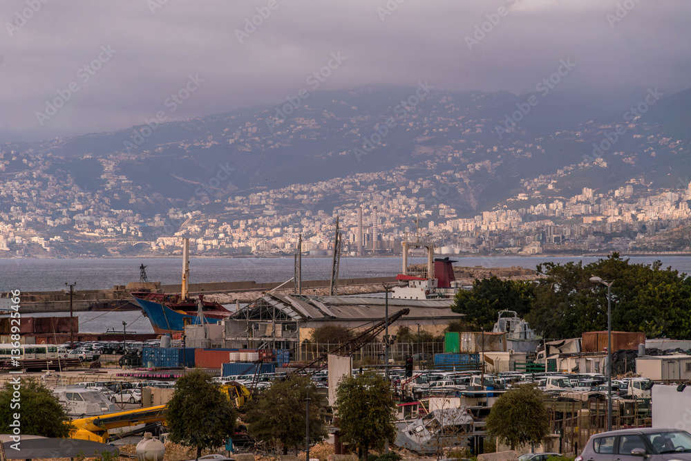 Obraz premium A panoramic view of Beirut port, Lebanon, with industrial port cranes, harbor activity, and a hilly landscape under an overcast sky.
