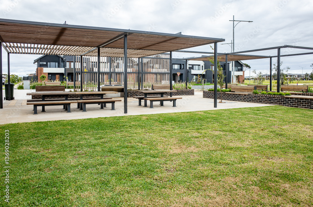Fototapeta premium A modern outdoor seating area with covered picnic tables,green lawn in a suburban park or community space.Minimalist modern pergola structures provide shade over the rest area in a suburb in Australia