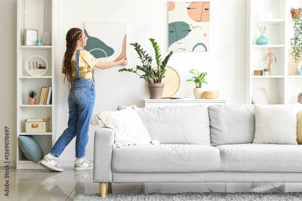 Young woman with dreadlocks hanging painting on wall at home Young woman with dreadlocks hanging painting on wall at home