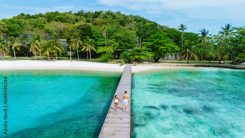 Couple strolls hand-in-hand along a wooden pier leading to the serene waters of Koh Kham Thailand, The vibrant turquoise sea meets the sandy shore, framed by palm trees and tropical foliage. Couple strolls hand-in-hand along a wooden pier leading to the serene waters of Koh Kham Thailand, The vibrant turquoise sea meets the sandy shore, framed by palm trees and tropical foliage.
