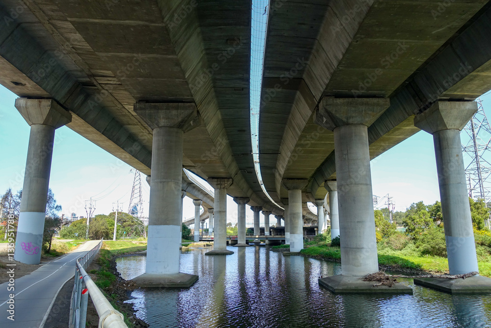Fototapeta premium Underside of a large concrete highway overpass or elevated roadway crossing over the Moonee Ponds Creek in Melbourne, Australia. Concept of urban infrastructure, waterway management, civil engineering