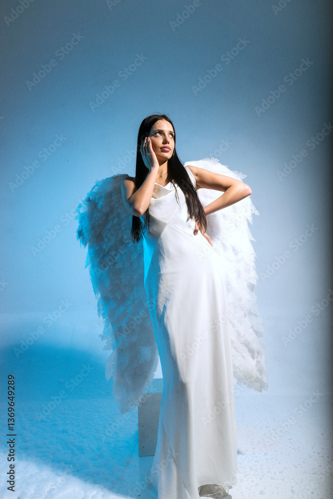 studio photo of blonde in white long dress and angel wings posing on blue background in studio studio photo of blonde in white long dress and angel wings posing on blue background in studio