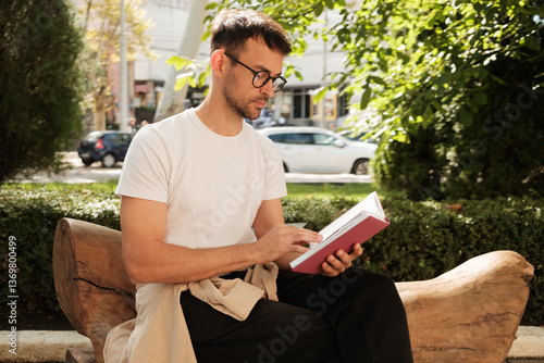 A man sits on a wooden bench in a sunny park, focused on reading a book. Surrounded by greenery, he enjoys a peaceful moment of relaxation and solitude.