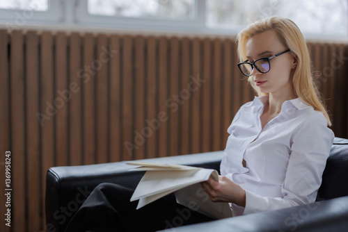 Woman in glasses reading documents while sitting on a couch in a cozy indoor setting during daylight hours
