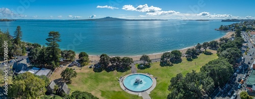 Aerial view of Auckland's Mission Bay, New Zealand, showing the beach, park with fountain, and Rangitoto Island in the distance. People are enjoying the sunny day. AUCKLAND, NZ