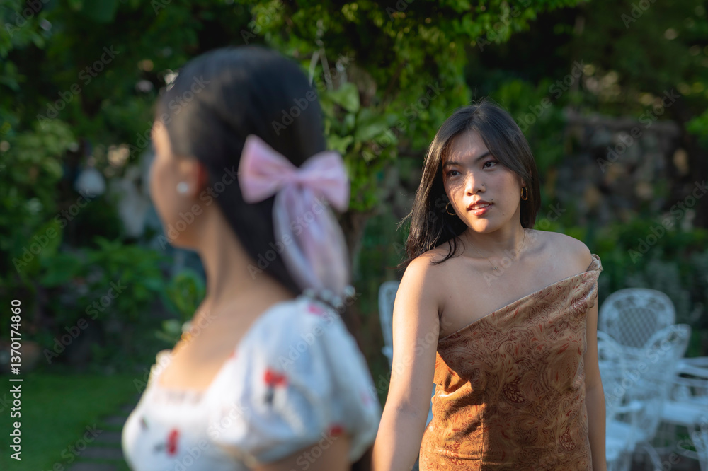 Naklejka premium Southeast Asian women in their early 20s walk hand in hand outdoors, one leading the other with a caring expression in a lush, sunlit garden