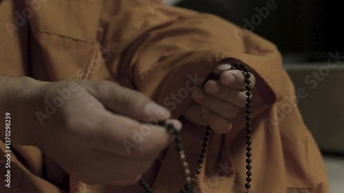 A close-up of the hands of a monk in an orange robe, who is fingering small brown rosaries during his meditation