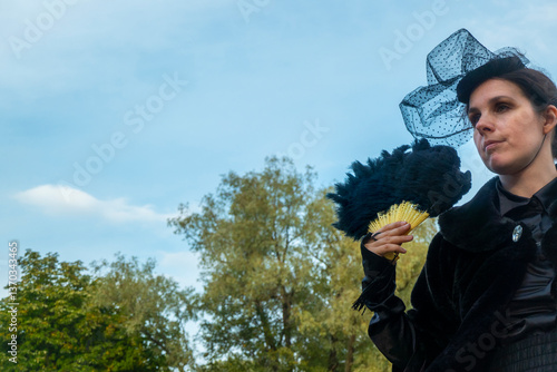 Obraz na płótnie A confident and sophisticated lady in a strict black suit of the 19th century posts with a fan against the backdrop of an park. w salonie