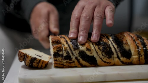 A man is cutting a poppy seed cake. The poppy seed cake is cut in half. The knife is on the left side of the bun.