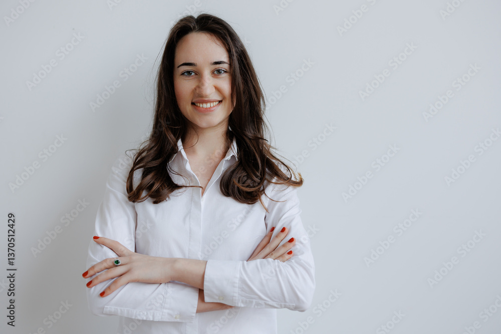Young woman smiling confidently with arms crossed in front of a neutral background during a casual indoor setting Young woman smiling confidently with arms crossed in front of a neutral background during a casual indoor setting