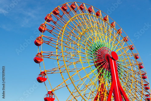 Obraz na płótnie A wide shot looking up at a rainbow-colored carnival wheel illuminated by a sunny day, against clear blue sky in the background w salonie
