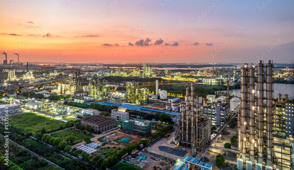 Obraz premium Aerial view of illuminated oil refinery at dusk with industrial zone waterfront and colorful sky background
