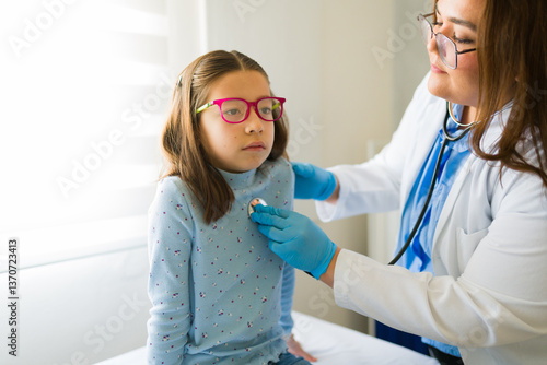 Schilderij op canvas Female pediatrician listening to girl's heart during medical checkup, wearing wh
