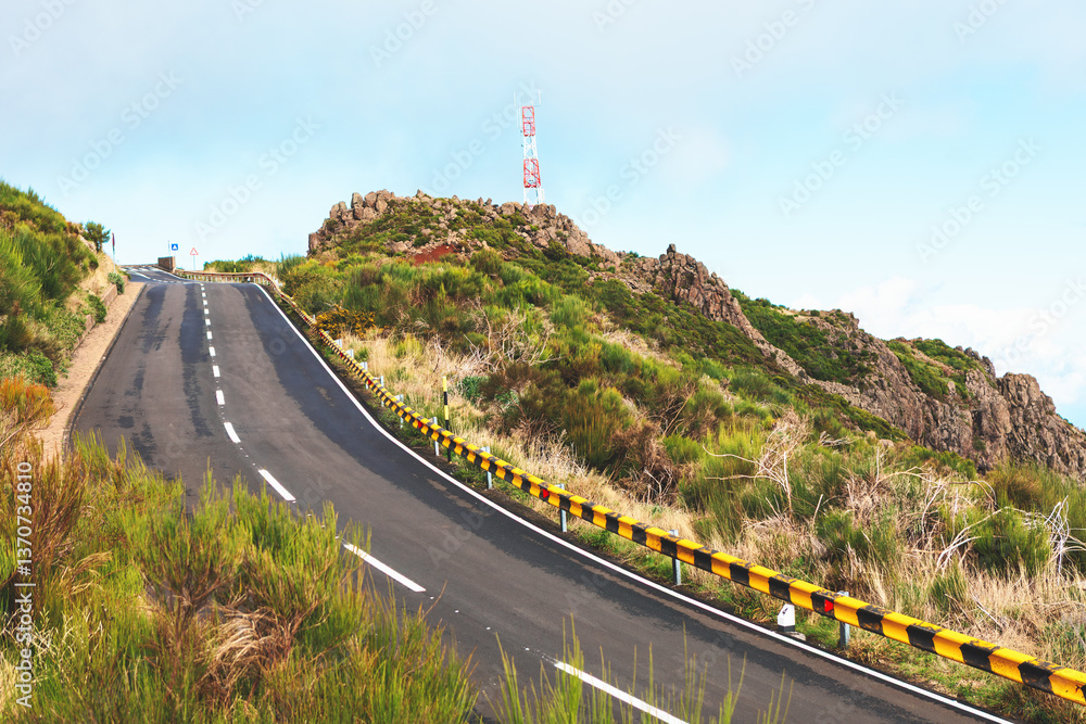 Winding road with striped guardrails curves through lush greenery, leading to atop a hill. Steep mountain road snakes its way upward. Asphalt road curves sharply, leading through mountainous terrain