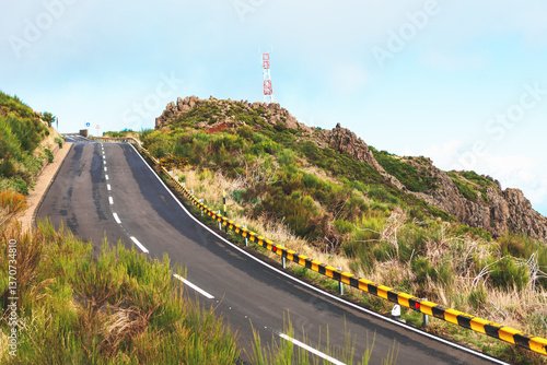 Obraz na płótnie Winding road with striped guardrails curves through lush greenery, leading to atop a hill. Steep mountain road snakes its way upward. Asphalt road curves sharply, leading through mountainous terrain w salonie