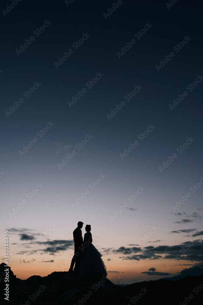 Silhouettes of the bride and groom standing on the mountain, holding hands at dusk Silhouettes of the bride and groom standing on the mountain, holding hands at dusk