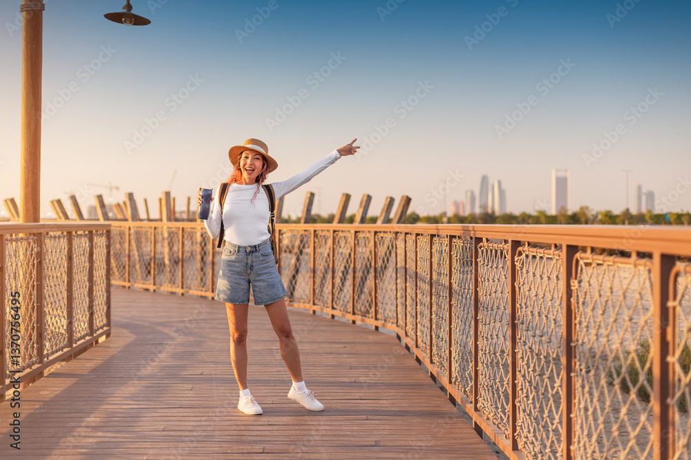 Young woman tourist pointing to cityscape in the background, enjoying the sunset while walking on a wooden boardwalk on Hudayriyat island in Abu Dhabi