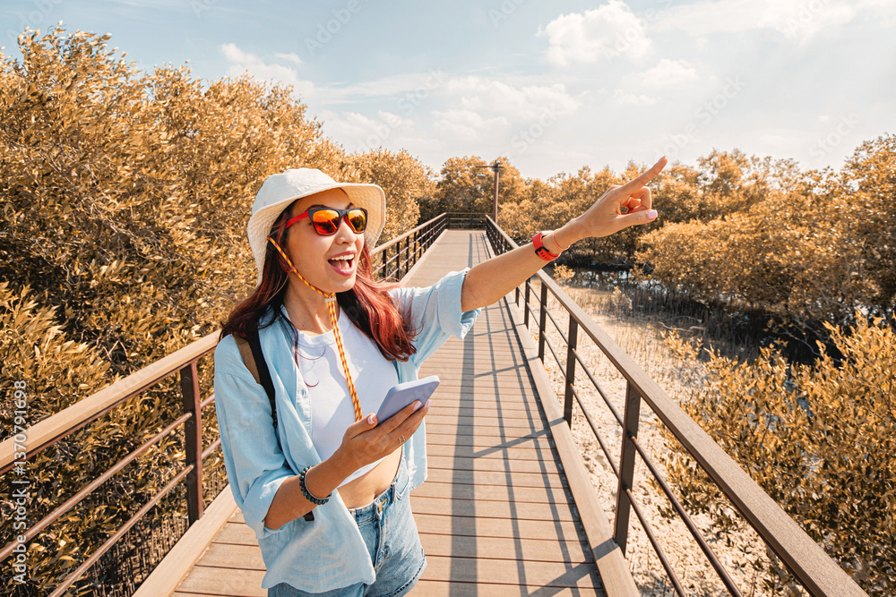 Excited woman in a sun hat and sunglasses pointing ahead while holding a smartphone on a wooden walkway through lush green mangroves
