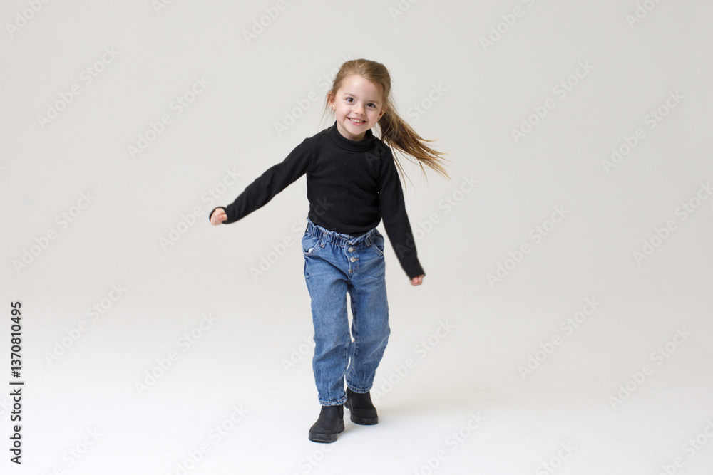 Happy little girl dancing with joy. style - Black sweater, blue jeans, and boots, smiling and moving energetically against a neutral background.