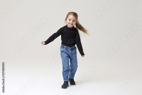 Obraz na płótnie Happy little girl dancing with joy. style - Black sweater, blue jeans, and boots, smiling and moving energetically against a neutral background. w salonie