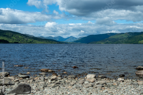 A scenic capture of Loch Katrine surrounded by majestic mountains and vivid greenery under a partly cloudy sky, located in the renowned Trossachs area of Scotland - UK