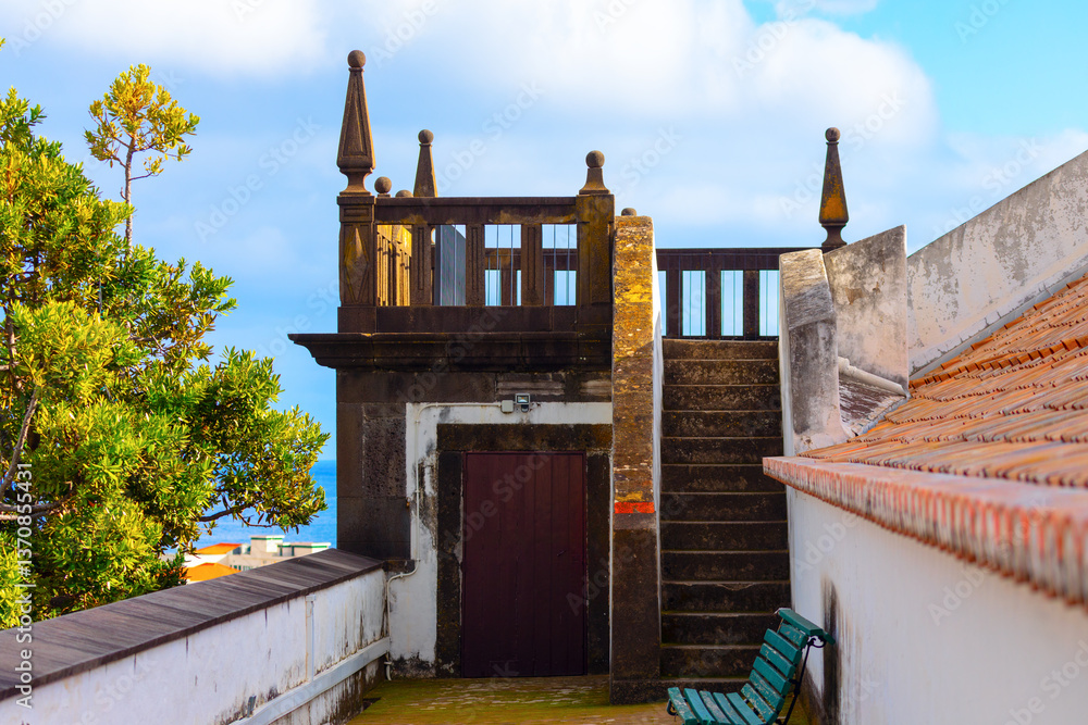 Church rooftop terrace with weathered stone balustrades and a narrow staircase leading to a scenic viewpoint. Green bench sits against a white wall, overlooking lush greenery and a distant ocean