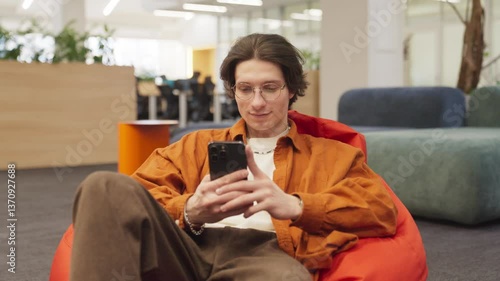Relaxed man wearing glasses and orange shirt sitting on bean bag in modern lounge while using smartphone. Scrolling through social media with focused expression. Enjoying break while engaging online.