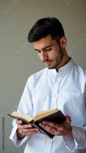 Young man reading a book indoors, focused on the pages, wearing a white shirt, natural light illuminating the scene, intellectual pursuit concept
