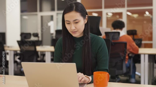 Focused Asian woman sitting in modern office. Typing on keyboard while working on laptop. Concentrating on task with serious expression. Completing assignment efficiently with full attention.