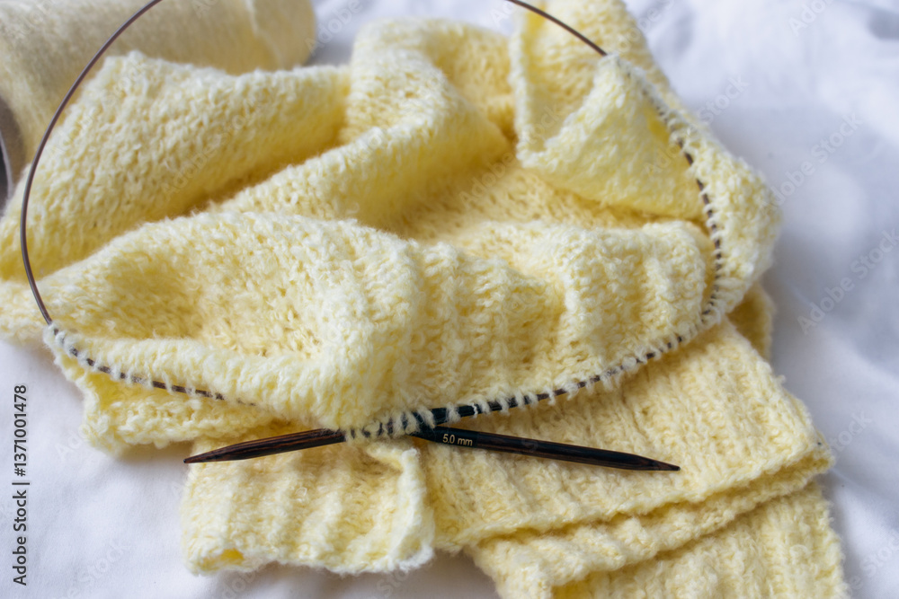 Yellow cotton knitting with knitting needles on white sheet. Soft yellow knitwear with circle needles. Knitting pattern background. Knitting lifestyle. Cozy hobby. Warm knitwear. Craft product concept