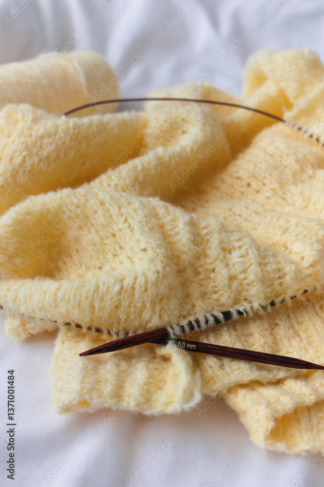 Yellow cotton knitting with knitting needles on white sheet. Soft yellow knitwear with circle needles. Knitting pattern background. Knitting lifestyle. Cozy hobby. Warm knitwear. Craft product concept