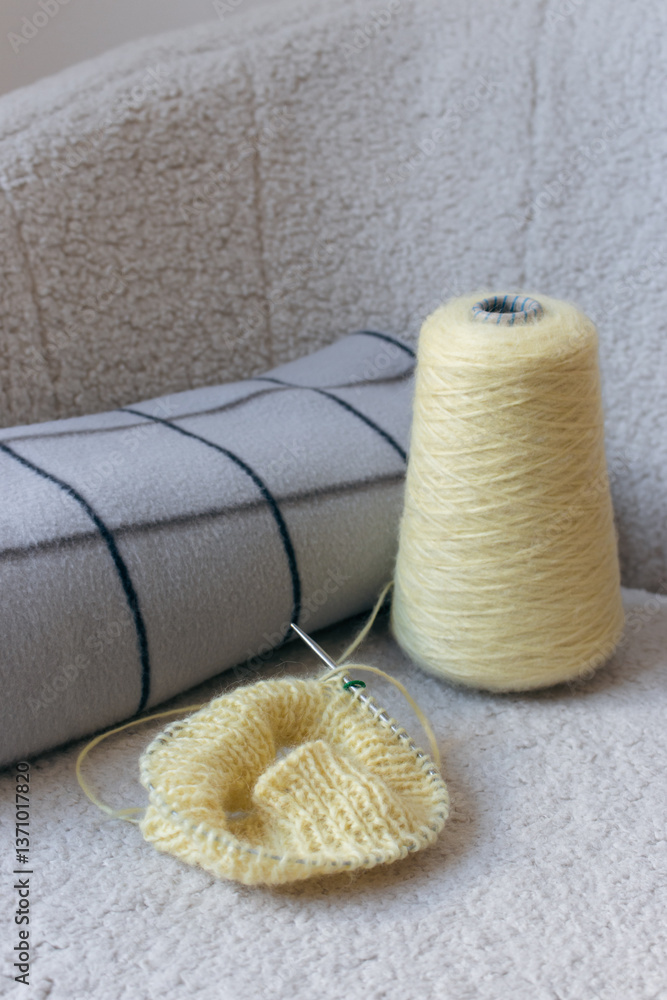 Yellow knitting with grey checkered plaid on sofa. Knitted fashion. Cotton knitting crochet with knitting needles and bobbin. Knitting hobby. Handmade knitted clothing. Cozy still life. Domestic life