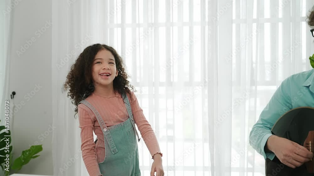 Happy schoolgirl with headphone standing and dancing to acoustic music while dad playing guitar. Smiling child move to music while enjoy to listening father song. Family recreation concept. Pedagogy. Happy schoolgirl with headphone standing and dancing to acoustic music while dad playing guitar. Smiling child move to music while enjoy to listening father song. Family recreation concept. Pedagogy.