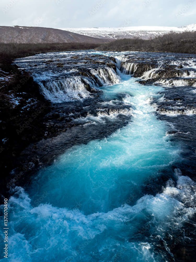 Bruarafoss Waterfall, Iceland: Turquoise glacial waters flow over rugged rocks, creating a mesmerizing cascade. Surrounded by winter landscape, it’s a natural wonder Bruarafoss Waterfall, Iceland: Turquoise glacial waters flow over rugged rocks, creating a mesmerizing cascade. Surrounded by winter landscape, it’s a natural wonder