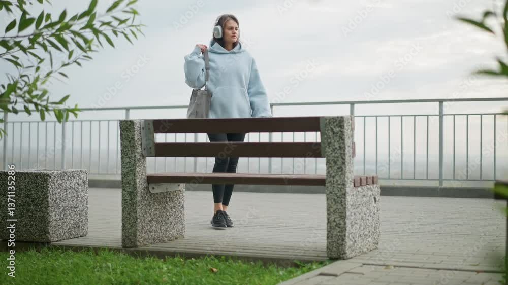 Lady walking toward concrete bench, carrying bag and wearing headphones, sitting carefully on bench in outdoor park, ideal for videos featuring relaxation, outdoor leisure, and tranquil moments Lady walking toward concrete bench, carrying bag and wearing headphones, sitting carefully on bench in outdoor park, ideal for videos featuring relaxation, outdoor leisure, and tranquil moments