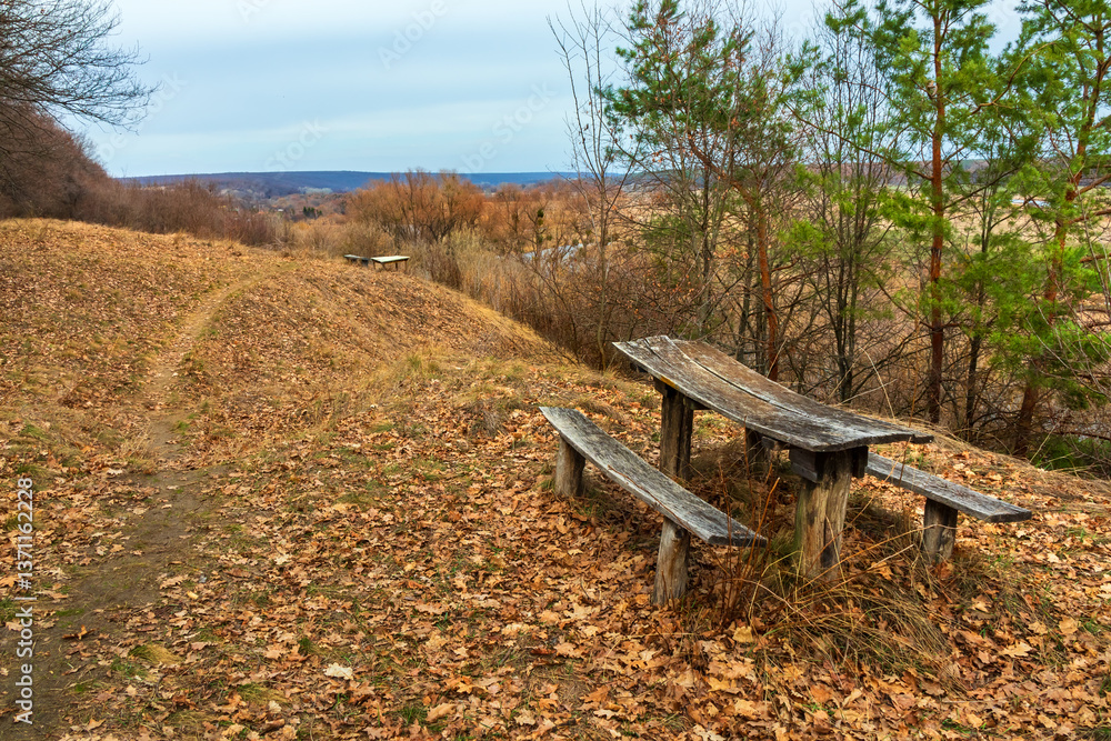 Fototapeta premium Old wooden table with benches near a hiking trail on a hill near a forest. Picturesque countryside landscape in Ukraine. Fallen autumn leaves on yellow dry grass