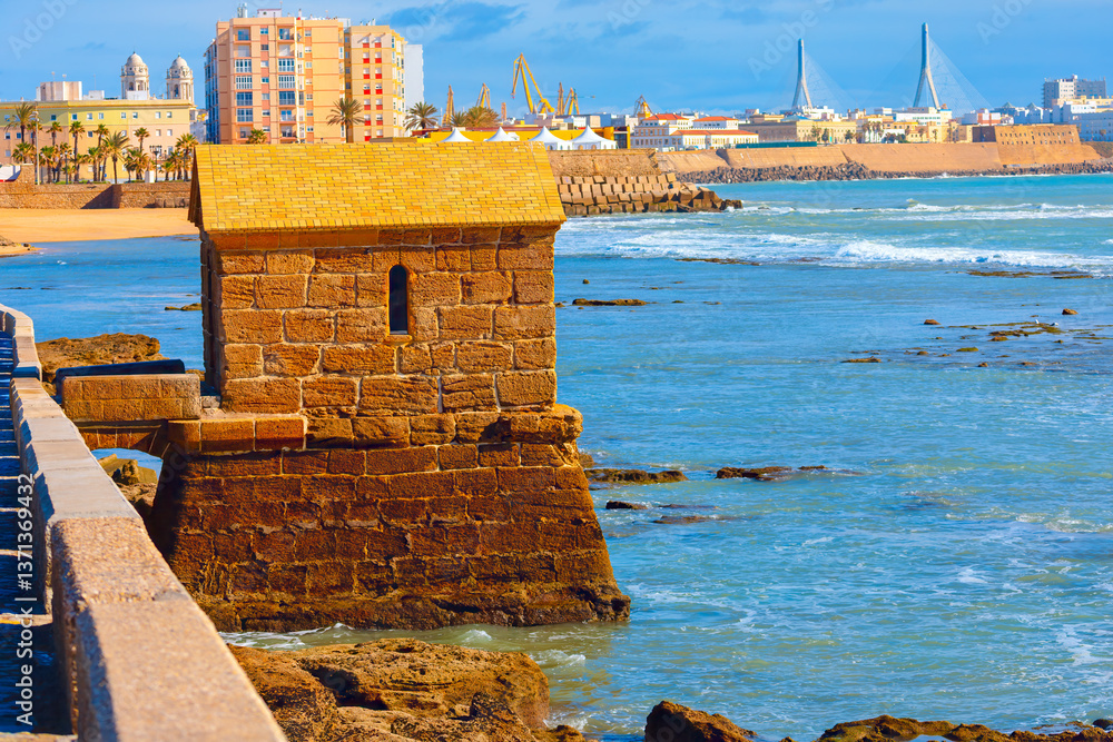 Historic fort stands at the edge of ocean in Cadiz, Spain, blending with rugged coastline. In background, modern buildings, sandy beach and striking cable-stayed bridge contrast with city heritage