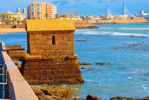 Obraz na płótnie Historic fort stands at the edge of ocean in Cadiz, Spain, blending with rugged coastline. In background, modern buildings, sandy beach and striking cable-stayed bridge contrast with city heritage w salonie