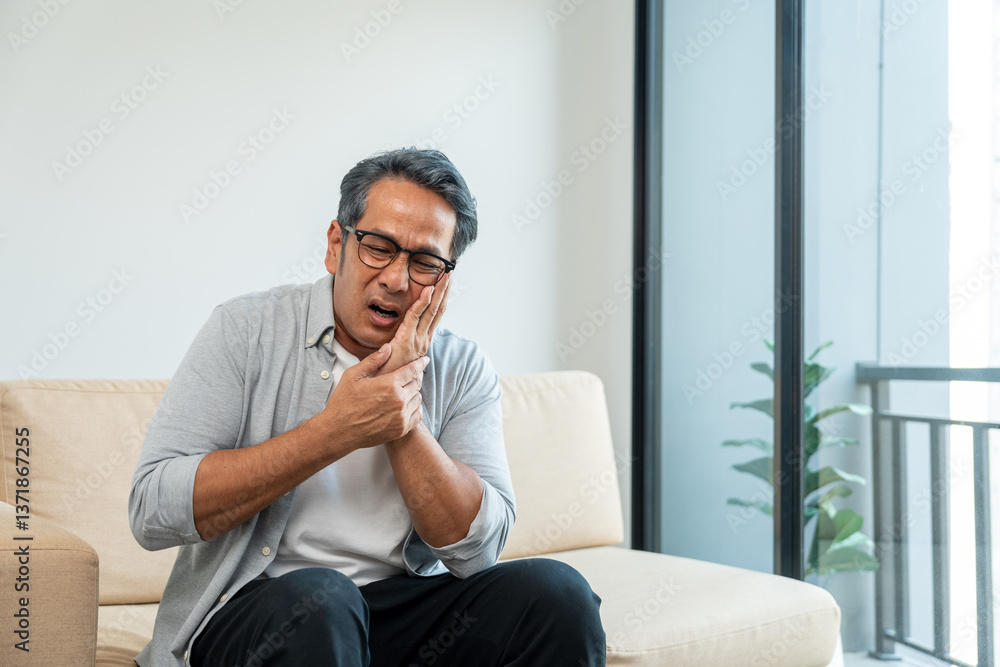 Fototapeta premium Old man senior puts his hand on his mouth and feels toothache because of tooth decay. Problems with teeth bad breath. Mature man unwell unhealthy sitting on sofa in living room