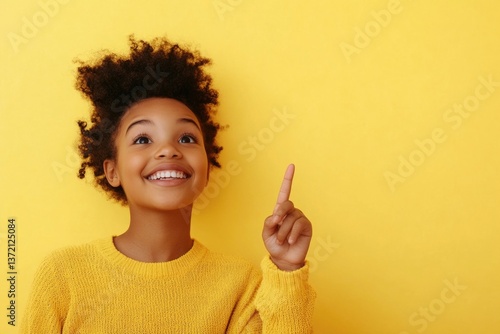 Child in bright yellow sweater smiling and pointing against vivid yellow wall during sunny day