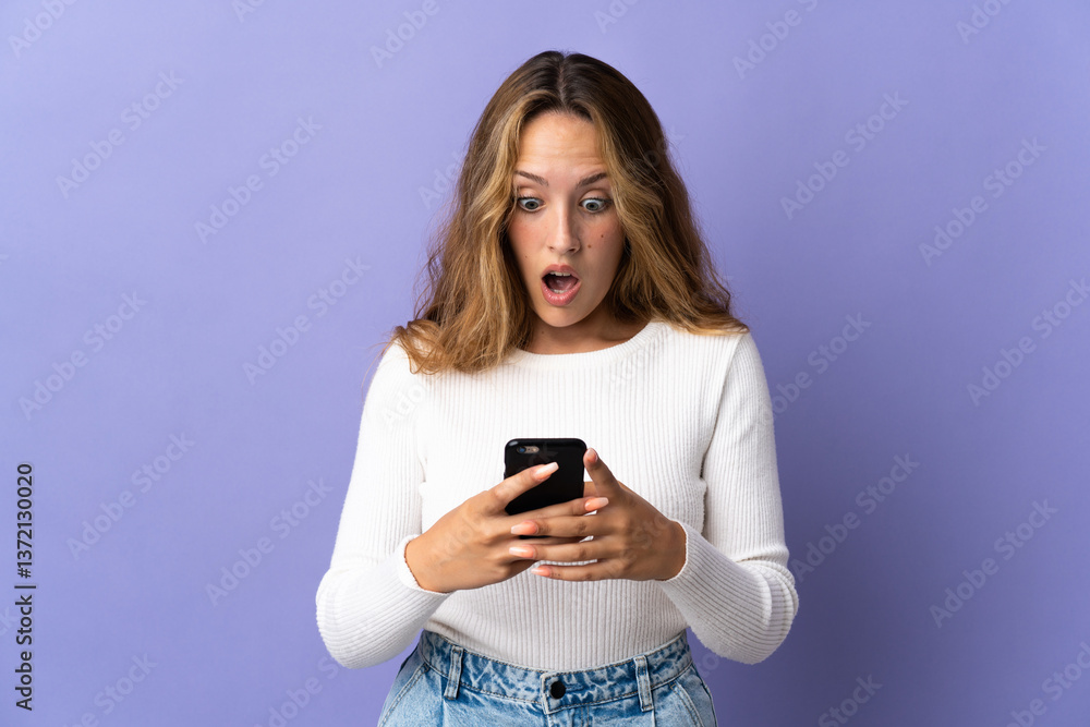 Fototapeta premium Young blonde woman isolated on purple background looking at the camera while using the mobile with surprised expression