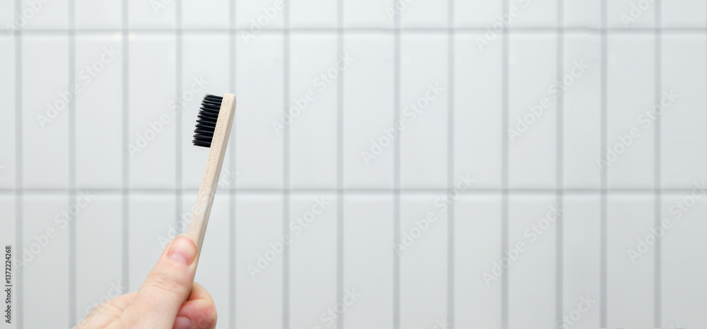 A wooden eco-friendly toothbrush held in a hand against a clean white tile background, emphasizing themes of sustainability, oral hygiene, and natural living. Copy space