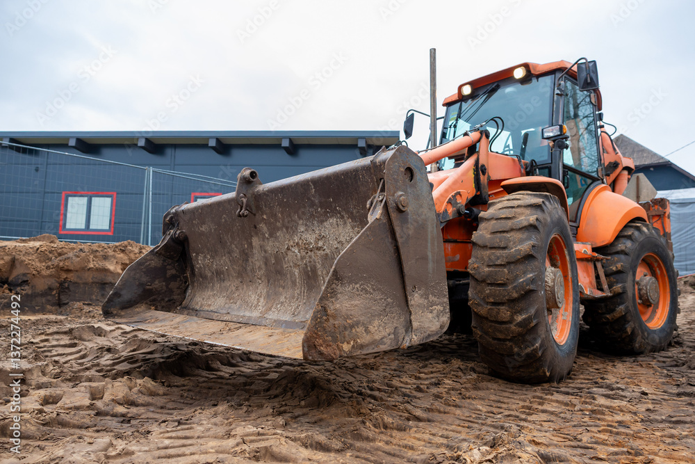 An orange front loader with a worn bucket sits on a dirt construction site. A blue building with red trimmed windows and a fence is in the background.