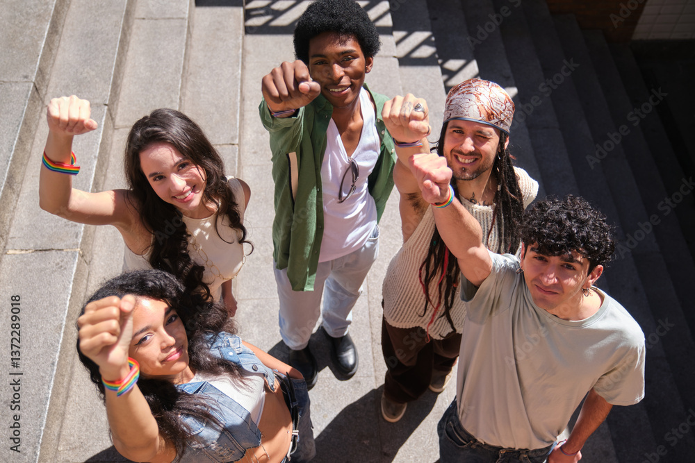 Group of diverse young people raising fists in a sign of protest and activism, wearing rainbow wristbands, standing on city steps