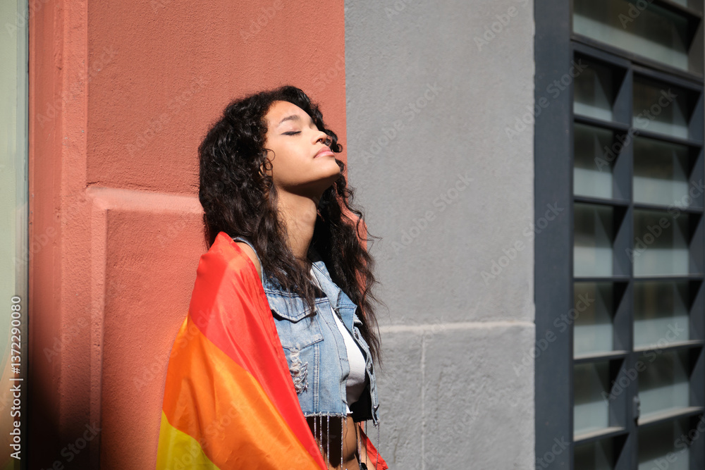 Young woman with LGBT rainbow flag enjoying sunlight against colorful wall in the city