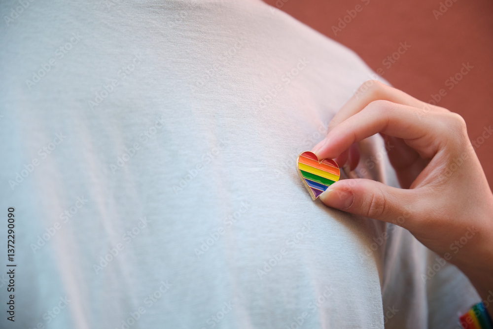 Close up of hand pinning rainbow heart to white t shirt, symbolizing support and celebration of LGBT pride